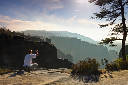 Man Takes photos with smart phone on the peak of rock empire. Dreamy fogy landscape, spring orange pink misty sunrise in a beautiful valley of Saxony Switzerland Park.の写真素材