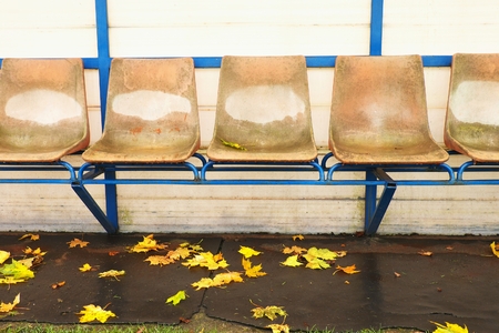 Old plastic seats on outdoor stadium players bench, chairs with worn paint below yellow roof.  Autumn leaves, end of football season.の写真素材
