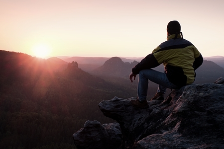 Rear view of male hiker in yellow black jacket sitting on the rocky peak while enjoying a daybreak above mountains valleyの写真素材