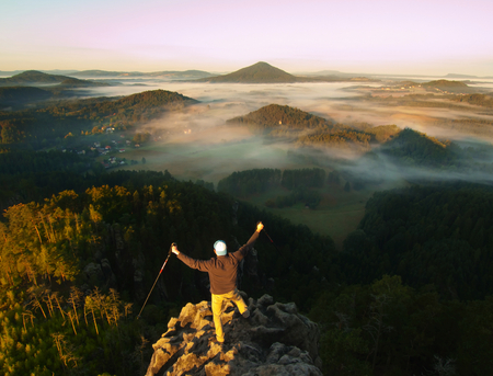 Happy tourist with poles in hand. Hiker with sports backpack stand on rocky view point above the misty valley. Sunny spring daybreak in the Rocky Mountains.の写真素材