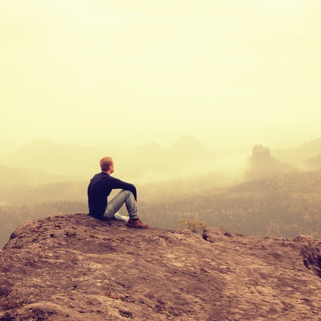 Young man in black shirt is sitting on cliff's edge and looking to misty valley bellowの写真素材