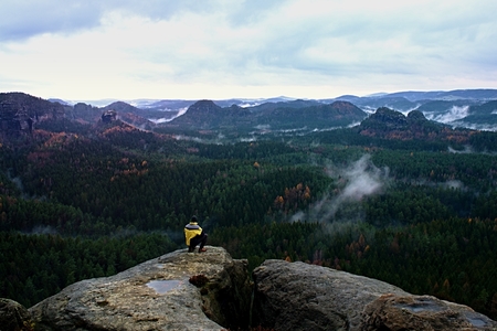 Rear view of male hiker in yellow black jacket on rocky peak while enjoying a daybreak above...