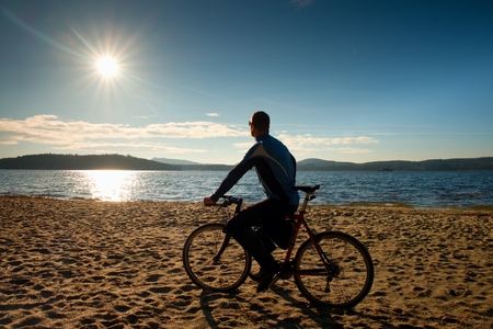 Young man cyclist sit on bike,  blue sky and sunset background on beach. End of season at lake and popular tourist resort..の写真素材
