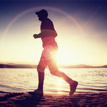 Running man in Sun rays on beach. Sportsman in baseball cap, jogging during the sunrise above sea level. Strong flare effect.の写真素材
