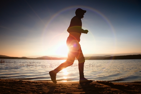 Running man in Sun rays on beach. Sportsman in baseball cap, jogging during the sunrise above sea level. Strong flare effect.の写真素材