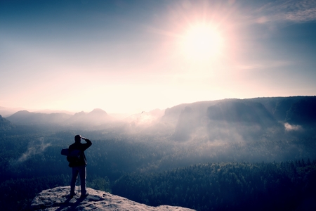 Tourist with backpack with sleeping pad and poles on a rocky peak. Sunny day, dreamy fogy valley belowの写真素材
