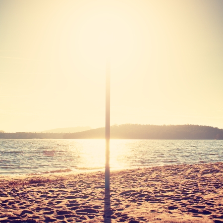 Sun hidden behind blue pole on sandy beach at sea. Forest hill on island in background. Vivid colorsの写真素材