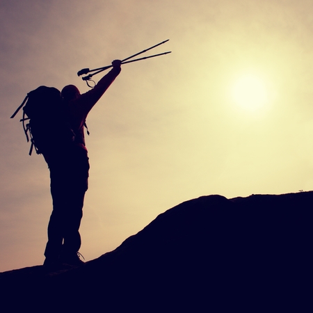 Silhouette of happy tourist with poles in hand above head. Sunny daybreak in mountains. Hiker with backpack sports stand on rocky view point above the misty valley.の写真素材
