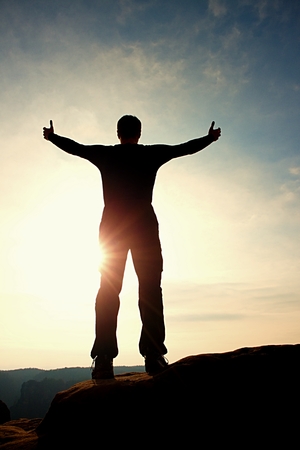 Gesture of triumph. Satisfy hiker in grey shirt and dark trousers. Tall man on the peak of cliff watching down to landscape.の写真素材