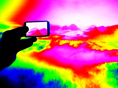 Fantastic infrared scan. Tourist hand takes photos by smartphone on view point and watching into misty spring valley.  Thermography colors.の写真素材