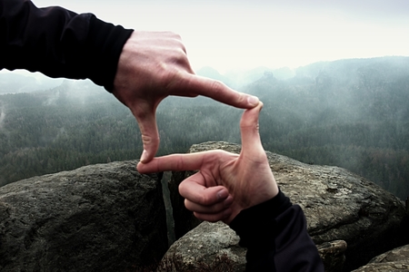 Close up of hands making frame gesture. Blue misty valley bellow rocky peak. Rainy spring in rocky mountains.の写真素材