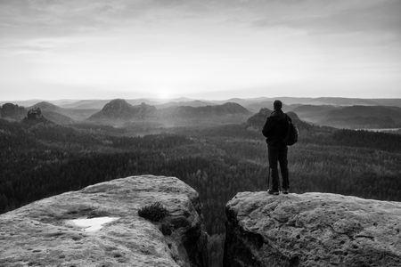 Tall tourist guide on rock with pole in hand. Hiker with sporty backpack stand on cliff above misty valley. Sunny spring daybreak in rocky mountains.の写真素材