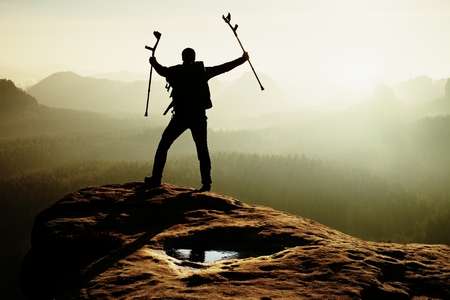 Hiker with broken leg in immobilizer and medicine pole above head achieved rocky summit. Deep misty valley bellow sharp edge. Silhouette of tourist with hand in air. Spring daybreakの写真素材