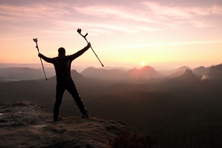 Hiker with  medicine crutch above head achieved mountain peak. Broken leg fixed in immobilizer Deep misty valley bellow silhouette of man with hand in air. Spring daybreakの写真素材