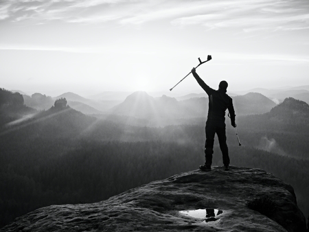 Tourist with forearm crutch above head achieved mountain peak. Hiker with broken leg in immobilizer and medicine  poles hold hand in air. Colorful misty valley bellow silhouette.の写真素材