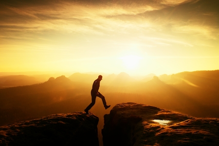Crazy hiker umping between rocks. Marvelous colorful daybreak in rocky mountains, heavy orange mist in deep valley. Miracle of natureの写真素材