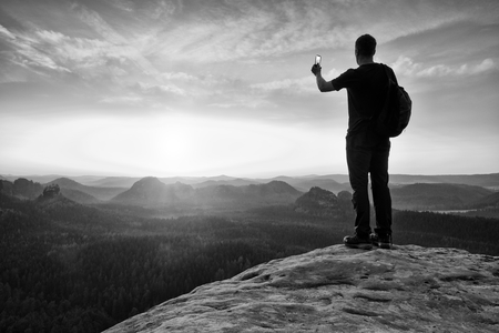 Tall backpacker with phone in hand. Sunny spring daybreak in rocky mountains. Hiker with backpack stand on rocky view point above misty valley.の写真素材