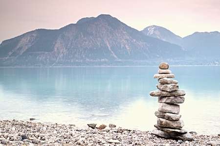Balanced stone pyramide on shore of blue water of mountain lake. Blue mountains in water level mirror. Children built pyramid from pebbles.  Poor lighting conditions.の写真素材