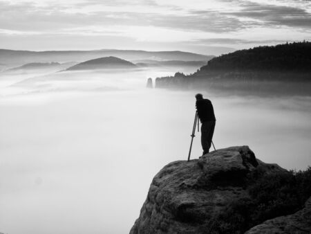 Professional photographer with tripod on cliff work. Dreamy fogy landscape, blue misty sunrise in a beautiful valley belowの写真素材
