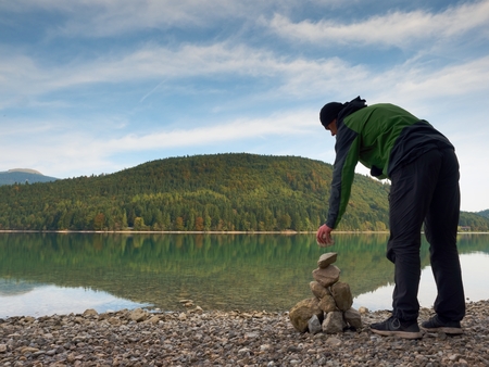 Man built pyramid from pebbles. Balanced stone pyramide on shore of blue water of mountain lake. Blue mountains in water level mirror. Poor lighting conditions.の写真素材