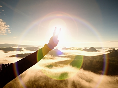 Flare. Strong lens defect, reflections. Man hand make victory symbol to Sun. Misty beautiful hills. Peaks of hills sticking out from creamy fogの写真素材