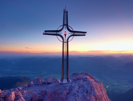 Steel crucifix at mountain peak in Alps. Sharp rocky summit, daybreak Sun in sky. Cross raised in...