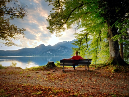 Man sit on wooden bench at mountain lake. Bank under beeches tree, mountains at horizon and in water mirror. Vintage toned photo.の写真素材