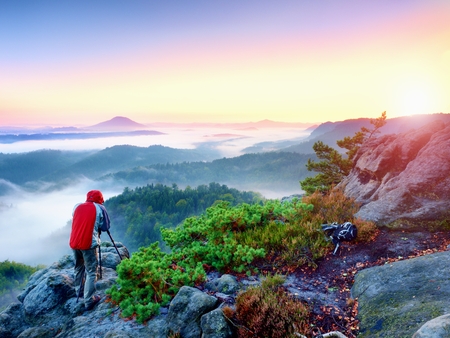 Photographer takes photos with mirror camera and tripod on peak of rock. Fall fogy landscape, spring orange pink misty sunrise in beautiful valley below.の写真素材