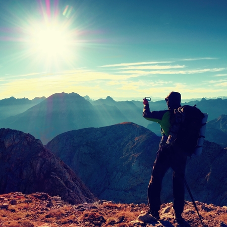 Experienced hiker takes phone photo. Man on Alps mountain peak.View to blue sky above deep foggy valley. Mountains increased from humidityの写真素材