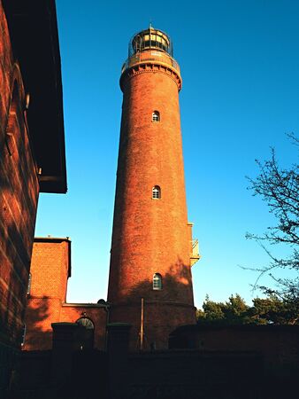 Shinning old lighthouse above houses and forest before sunset. Tower illuminated with strong warning light. Lighthouse built from red bricks, gallery with iron handrail around glass cover of spotlight.の写真素材
