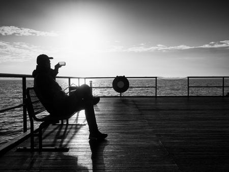 Tourist sit on steel bench on mole and take selfie.  Alone man enjoy misty morning at sea. Smooth water level. Vivid and strong vignetting effect.. の写真素材