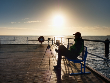 Hobby photograph sit on the bench at camera on tripod. Wooden board quay, sunny morning at sea. Smooth water level. Vivid and strong vignetting effect.の写真素材