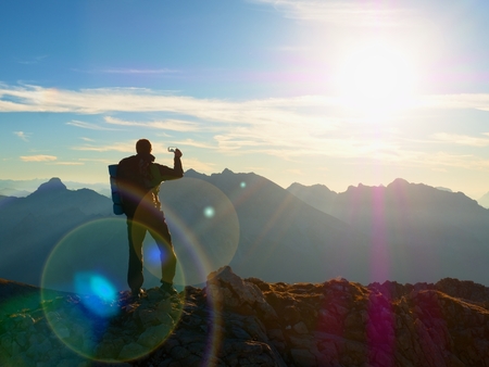 Lens flare light, strong defect. Hiker takes selfie photo. Man with big backpack and poles walk on Alpine mountain. Sun above horizon blue foggy valley. Mountains increased from humidityの写真素材