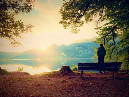 Man in green outdoor windcheater walk at lake bank.  Empty wooden bench, tree stump at mountain lake. Bank under beeches tree, mountains at horizon and in water mirrorの写真素材