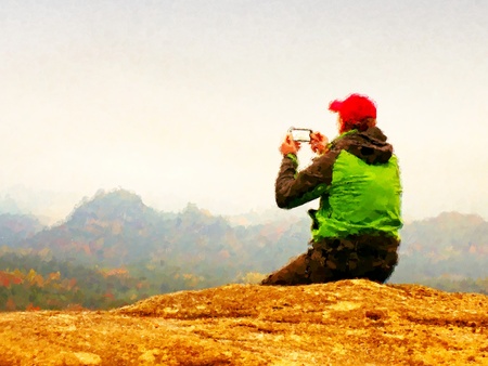 Watercolor paint effect. Man takes photos with smart phone on peak of rock empire. Melancholy fogy landscape, spring orange pink mistの写真素材