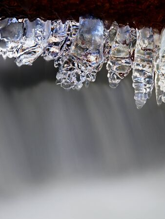 Long icicles hang above dark freeze  water of mountain stream. Winter season at river, thin icicles are hanging on fallen trunk above milky water level.の写真素材