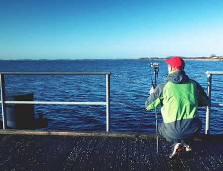 Tall nature photographer taking picture at tripod on the wooden pier in wharf at sunset time.の写真素材