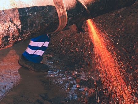 Worker staff is cutting big metal tube with hand grinder. Burning parks are flying down to wet clay....
