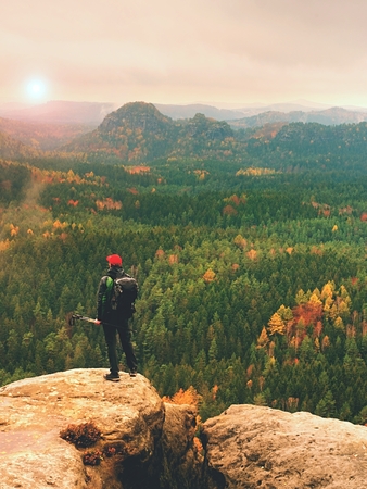 Tall hiker man with red cap,  poles in hand and with big backpack stand on rock. Misty spring...