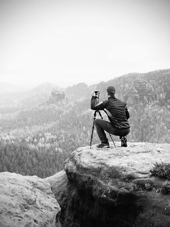 Outdoor photographer with tripod and camera on a rock thinking. Creamy autumnal mist in valley bellow. Dreamy rocky mountains. Misty sunrise in a beautiful valleyの写真素材