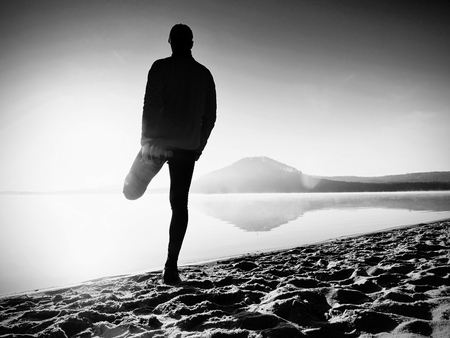 Man exercising on the beach.  Silhouette of active man exercising  and stretching on the lake beach at sunrise. Healthy lifestyle. Alone young fitness man exercise  at morning beachの写真素材