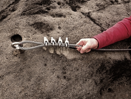 Rock climber hand hold on steel twisted rope at steel bolt eye anchored in rock. Tourist path via ferrata. The arm in red sporty jacket.の写真素材