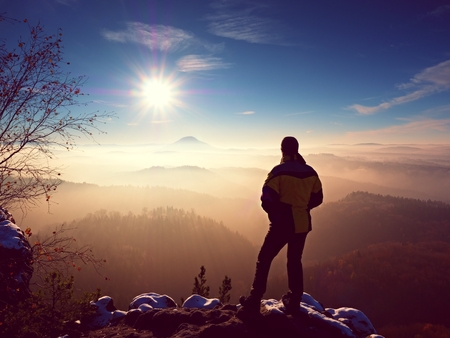 Tall hiker in black on rocky peak watch the world . Heavy orange mist bellow in valley. Wonderful breathtaking daybreak in mountains.の写真素材