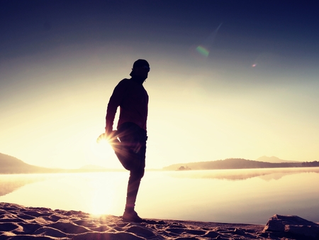 Silhouette of active man exercising  and stretching on the lake beach at sunrise. Healthy lifestyle. Alone young fitness man exercise  at morning beachの写真素材