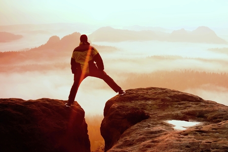Hiker in black is jumping between the rocky peaks. Wonderful daybreak in rocky mountains, heavy orange mist in deep valley.の写真素材