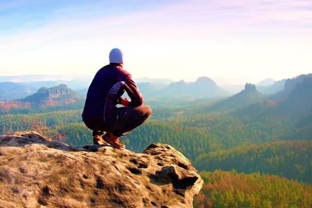 Young man in black sportswear is sitting on cliff's edge and looking to misty valley bellowの写真素材