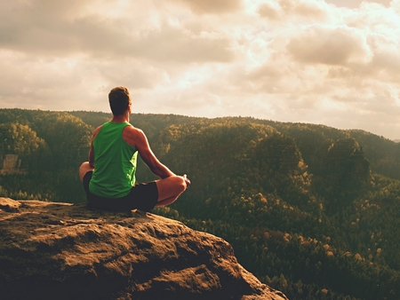 Man meditating in Lotus Pose on rocky cliff. Sportsman practicing Yoga on stone edge above landscapeの写真素材