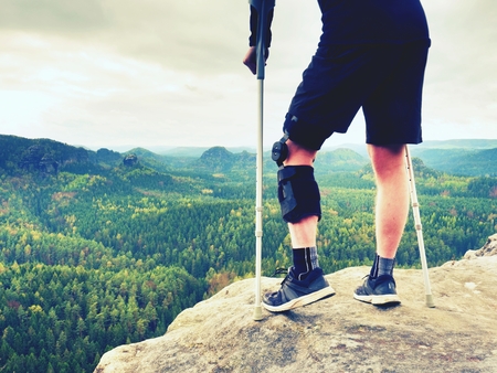 Tired hurt tourist with medicine crutches. Man with  broken leg in knee brace features resting on  exposed rocky summit. Valley bellow sitting man in black sweatshirt and red baseball cap. Sharp sandstone edge.の写真素材