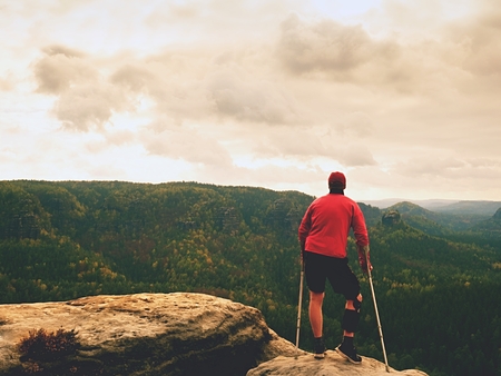 Man with forearm crutch. Hiker finaly achieved mountain peak. Tourist with broken leg in immobilizer hold medicine  poles.の写真素材