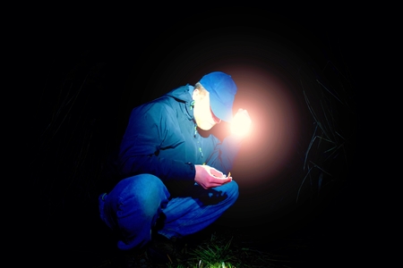 Adult man in blue wearing sit and looking for something in wet grass with light in hand , scary or fairytale nightの写真素材
